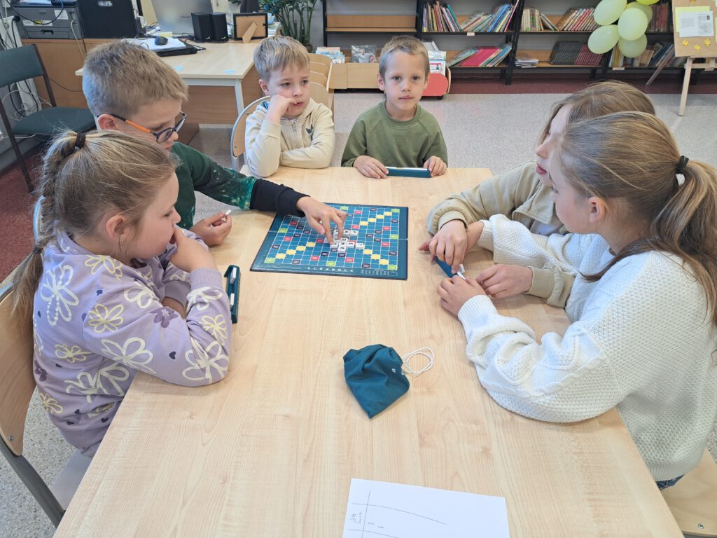 XXII Ogólnopolski Tydzień Bibliotek pod hasłem „Biblioteka. Lubię tu być". Przedstawia uczniów siedzących przy ławce i grających w Scrabble. Obok planszy leży zielony worek z literami. W tle książki na regałach.