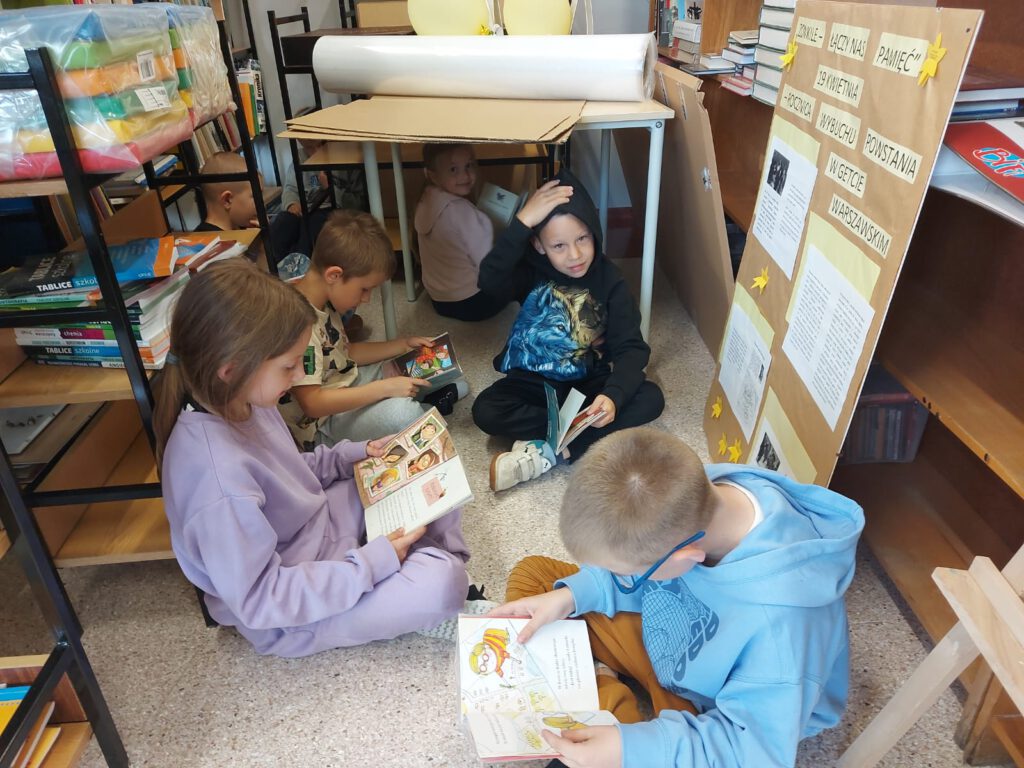 XXII Ogólnopolski Tydzień Bibliotek pod hasłem „Biblioteka. Lubię tu być”. Przedstawia dzieci, którzy czytają na podłodze w bibliotece książki. Wokół dzieci są regały, stoliki oraz sztalugi z gazetkami.