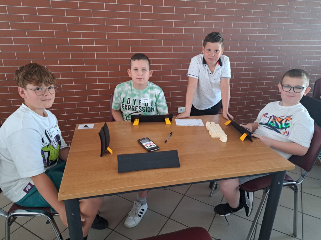 Międzyszkolny Turniej Rummikub w Radwanicach. Zdjęcie przedstawia chłopców z 4 klasy ubranych w kolorowe koszulki siedzących przy stole podczas Turnieju Rummikub. Na stole są podkładki do gry i kafelki z liczbami do gry.