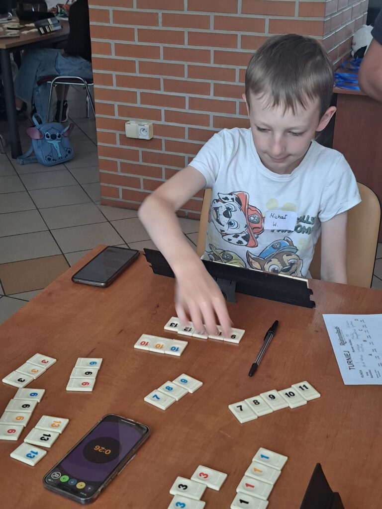 Międzyszkolny Turniej Rummikub w Radwanicach. Przedstawia ucznia podczas gry. Układa kafelki do sekwencji, czarne cyfry od 9 do 13.