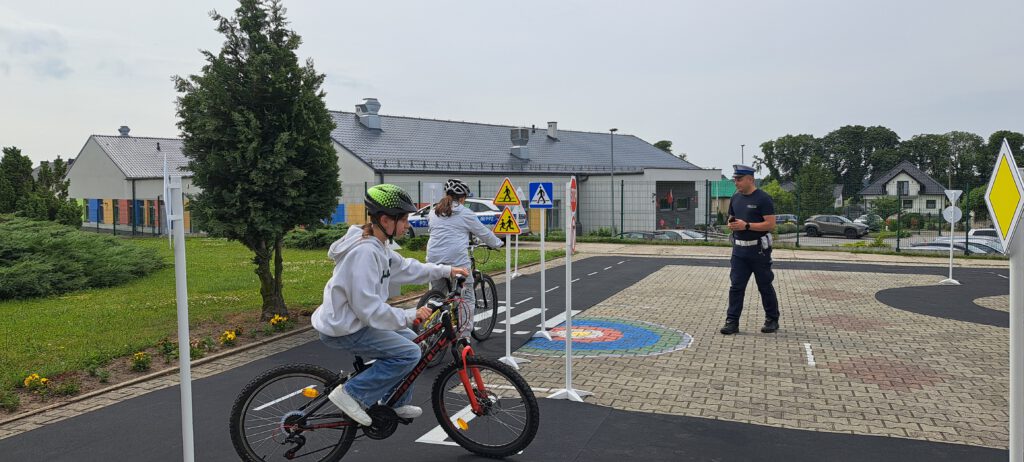 Egzamin na kartę rowerową. Na zdjęciu policjant egzaminuje dwie uczennice jadące rowerem po wyznaczonej trasie,