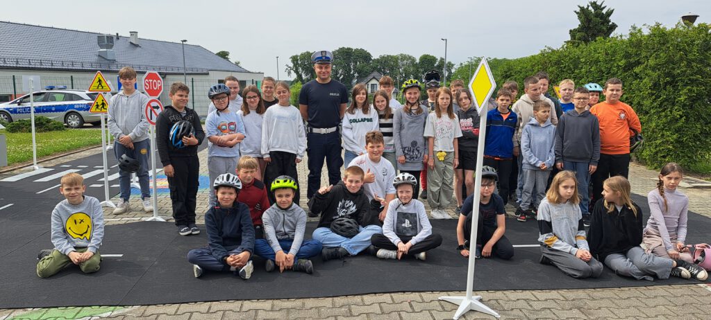 Egzamin na kartę rowerową. Na zdjęciu uczniowie klas czwartej a i b pozują w kaskach do zdjęcia grupowego z policjantem. Po prawej stronie znajduje się zielony żywopłot, po lewej widać budynek i radiowóz policyjny.