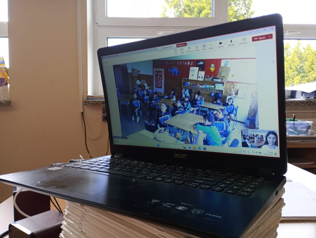 Na zdjęciu jest laptop, na którego monitorze widać połączenie online z siostrzaną szkołą z Hiszpanii.
Uczniowie z Hiszpanii siedzą w swojej klasie szkolnej. Ubrani są w niebiesko-granatowo-białe mundurki.