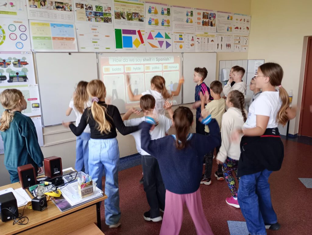 Dzieci wykonują ćwiczenia, które powiązane są z właściwym słówkiem w języku hiszpańskim, angielskim, polskim, duńskim
i ukraińskim. Ustawione są przed tablicą, na której wyświetlane są pytania.