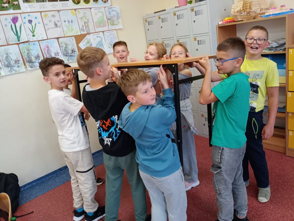 Na zdjęciu widać dziewięcioro dzieci, które są w klasie. Wykonują zadanie polegające na wspólnym podniesienu
palcami dłoni stolika.