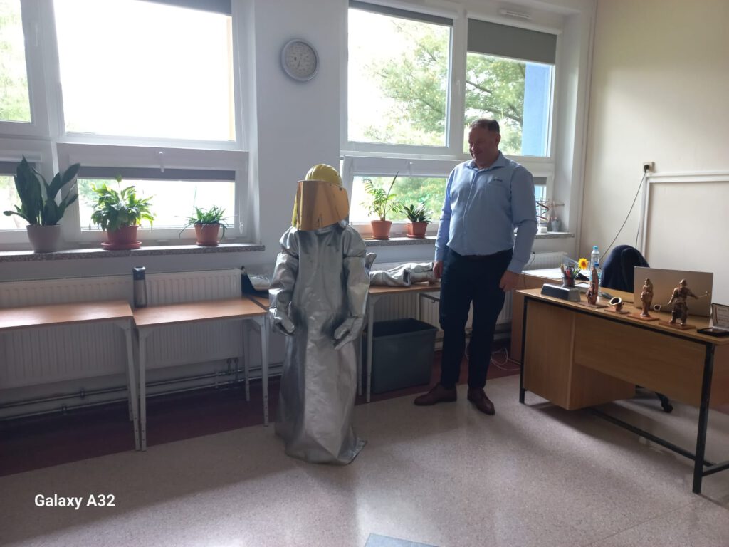Wizerunek prowadzącego warsztaty (hutnika) i ucznia ubranego w strój obowiązujący podczas wykonywania pracy w hucie przy bardzo wysokiej temperaturze.