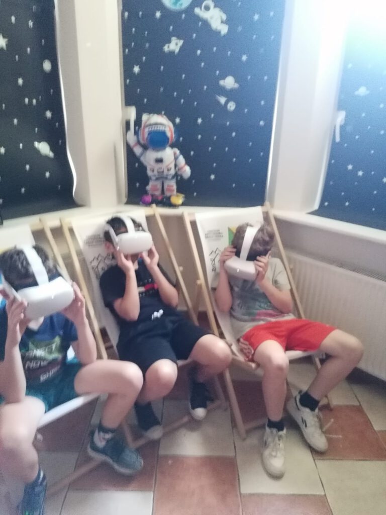 Na zdjęciu uczniowie siedzą na drewnianych leżaczkach i oglądają kosmos przez specjalne okulary.