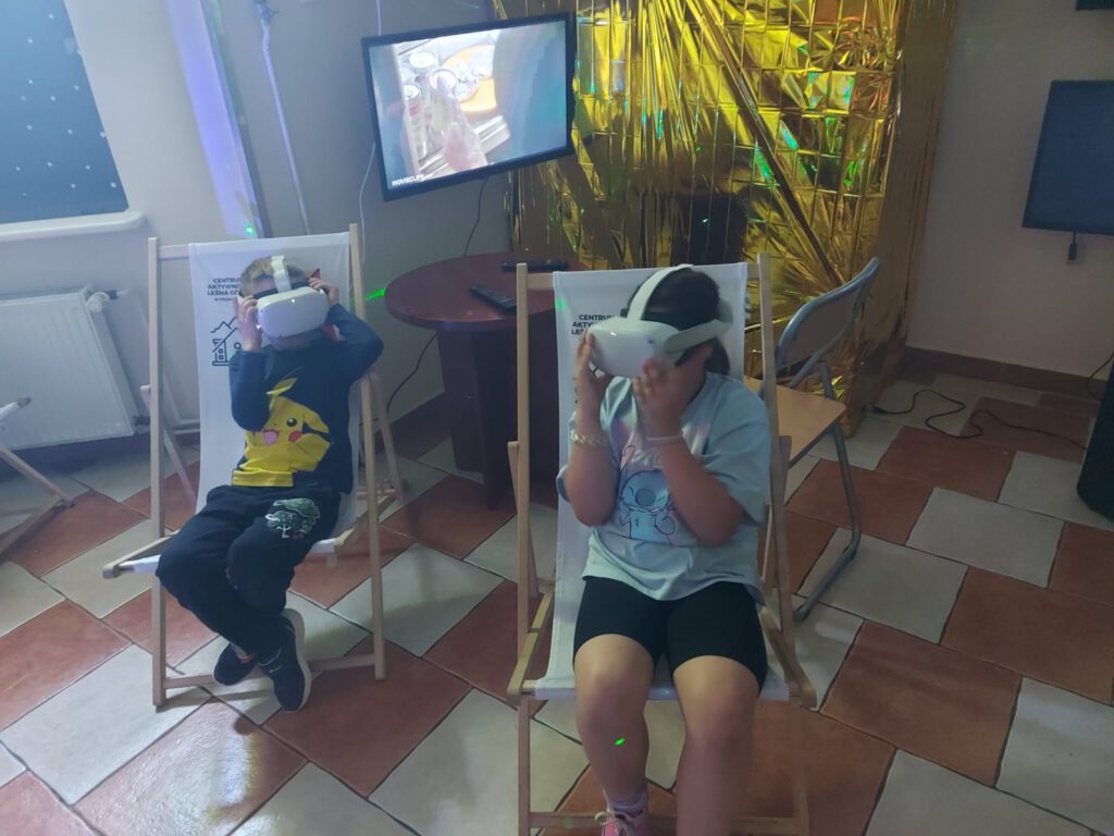 Na zdjęciu uczniowie siedzą na drewnianych leżaczkach i oglądają kosmos przez specjalne okulary.