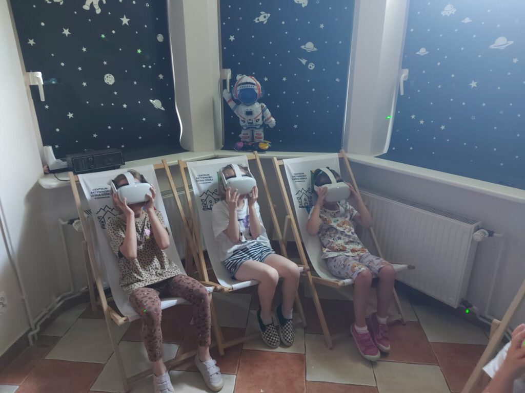 Na zdjęciu uczniowie siedzą na drewnianych leżaczkach i oglądają kosmos przez specjalne okulary.