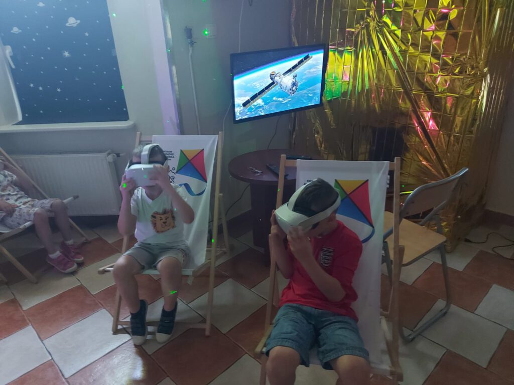 Na zdjęciu uczniowie siedzą na drewnianych leżaczkach i oglądają kosmos przez specjalne okulary.