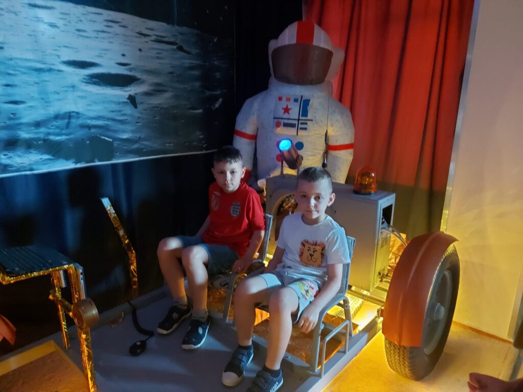 Dwóch chłopców siedzi w kosmicznym łaziku, w tle stoi nadmuchany strój astronauty.