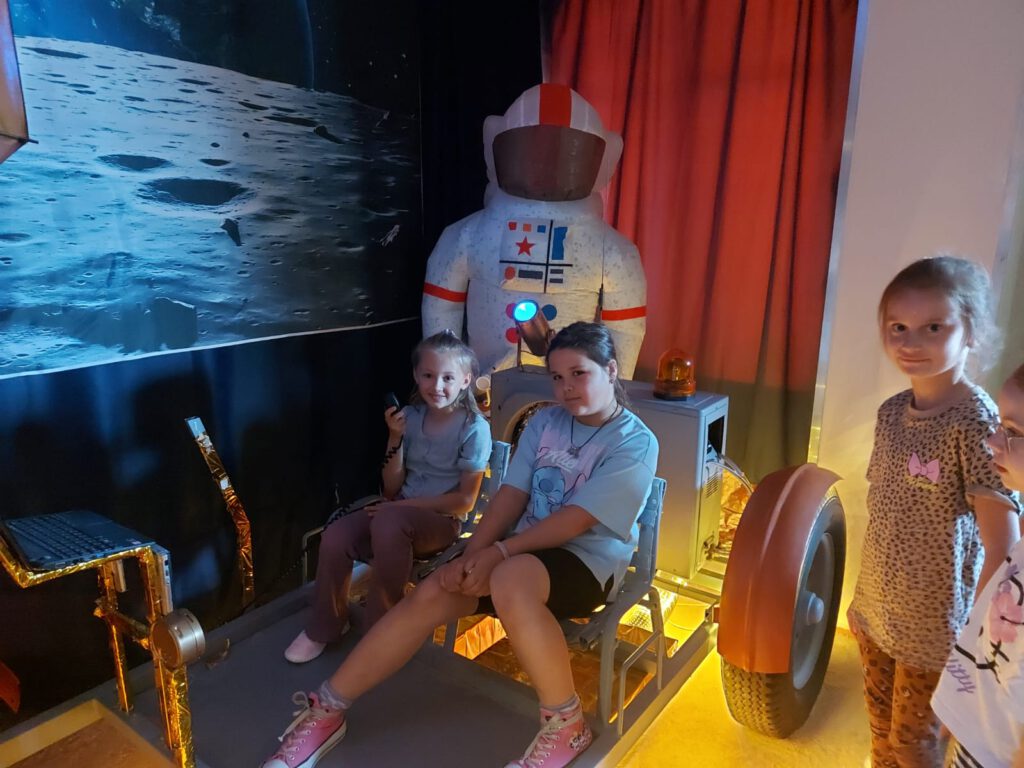 Dwie dziewczynki siedzą w kosmicznym łaziku, obok stoi inna dziewczynka, w tle stoi nadmuchany strój astronauty.