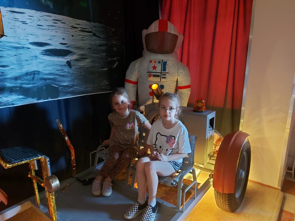 Dwie dziewczynki siedzą w kosmicznym łaziku, w tle stoi nadmuchany strój astronauty.