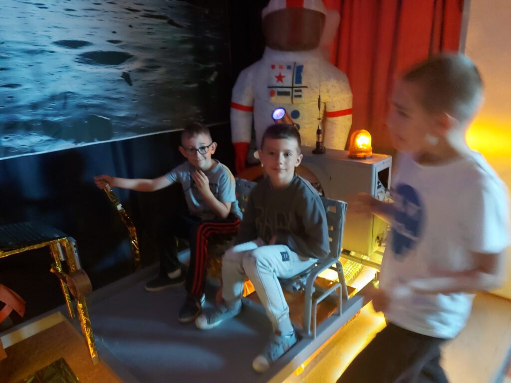Dwóch chłopców siedzi w kosmicznym łaziku, w tle stoi nadmuchany strój astronauty.