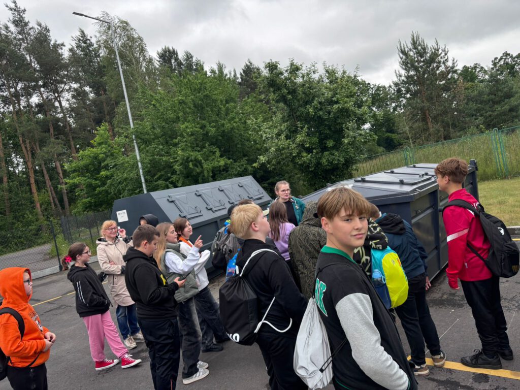 Projekt edukacyjny ,,Jestem w pSzoku”. Na zdjęciu uczniowie stoją przy kontenerach na śmieci i słuchają wykładu pani pracującej w PSZOKu.