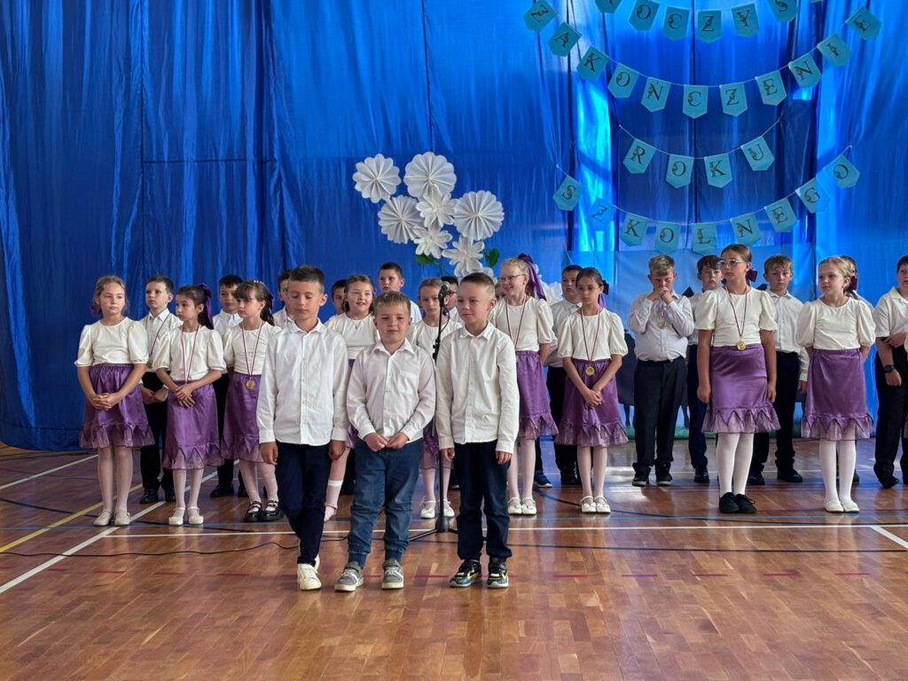 Pożegnanie uczniów klas trzecich. Na zdjęciu stoją uczniowie klas trzecich na tle granatowej kotary z napisem uroczyste zakończenie roku szkolnego. Dziewczynki ubrane są w białe bluzki i fioletowe spódniczki. Za dziewczynkami stoją chłopcy ubrani w białe koszule i ciemne spodnie. na pierwszym planie stoi trzech uczniów, którzy będą odbierać nagrody.