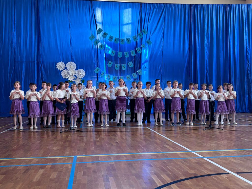 Pożegnanie uczniów klas trzecich. Na zdjęciu stoją uczniowie klas trzecich na tle granatowej kotary z napisem uroczyste zakończenie roku szkolnego. Dziewczynki ubrane są w białe bluzki i fioletowe spódniczki. Za dziewczynkami stoją chłopcy ubrani w białe koszule i ciemne spodnie.