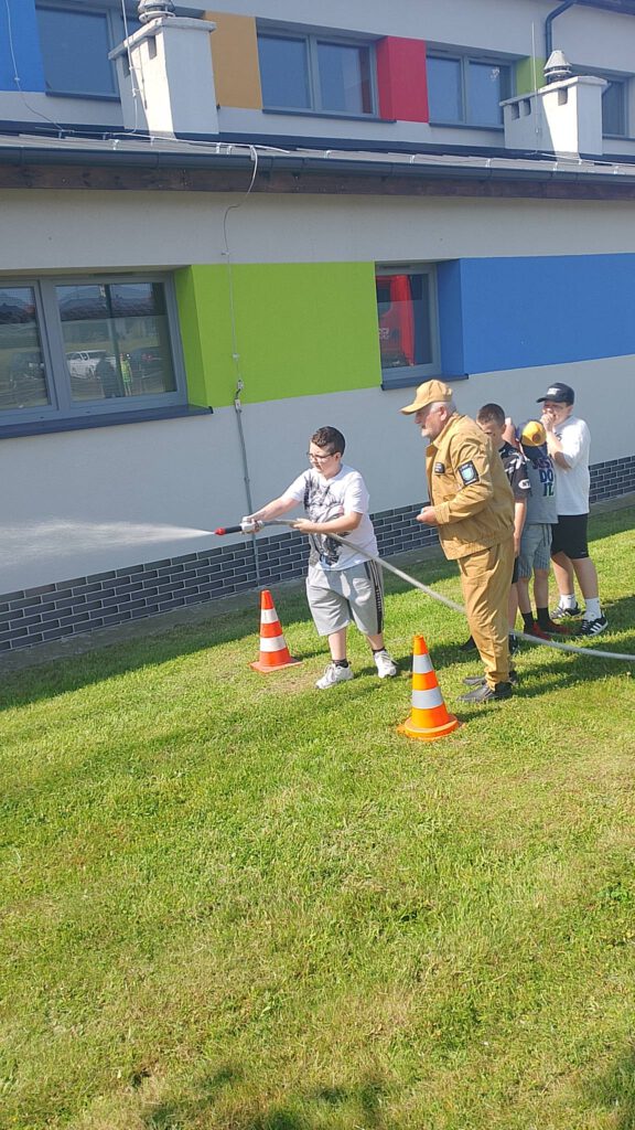 Piknik "Powitanie lata". Na zdjęciu uczeń trzymając armatkę wodną próbuje strącić piłkę. Przy nim stoi strażak i inni uczniowie.