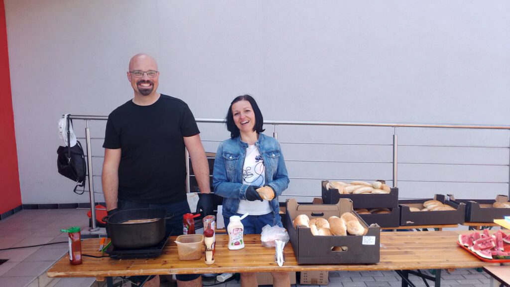 Piknik "Powitanie lata". Na zdjęciu dwoje rodziców stoi przy stoisku z hot dogami. Oboje są bardzo uśmiechnięci.