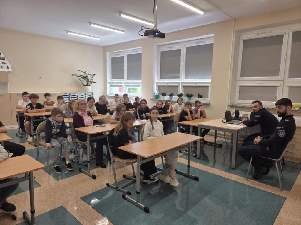 Spotkanie z policjantami na temat bezpieczeństwa. Na zdjęciu uczniowie klas 5a  i 5b siedzą na krzesełkach w klasie  w klasie  i słuchają policjantów.