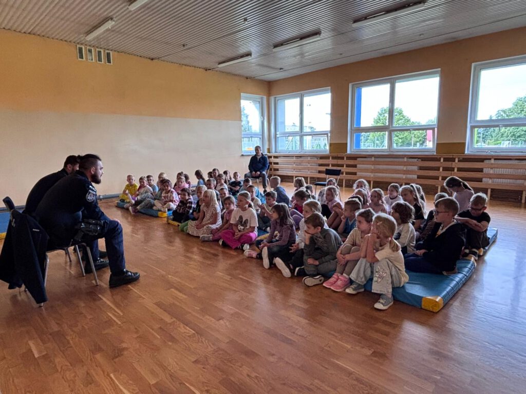 Spotkanie z policjantami na temat bezpieczeństwa. Na zdjęciu uczniowie kas 1 a, 1 b i 1 c  siedzą  na podłodze w salce gimnastycznej i słuchają policjantów.