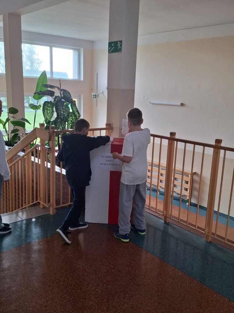 Wybory do Samorządu Uczniowskiego. Na zdjęciu dwóch uczniów wrzuca karty głosowania do urny wyborczej.