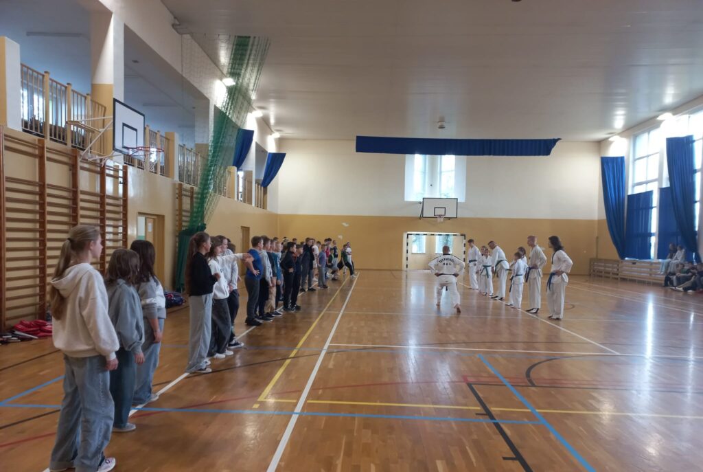 Europejski Dzień Sportu Szkolnego. Na zdjęciu trenerzy taekwondo pokazują uczniom ustawionym w szeregu elementy rozgrzewki: wykroki rozciągające.
