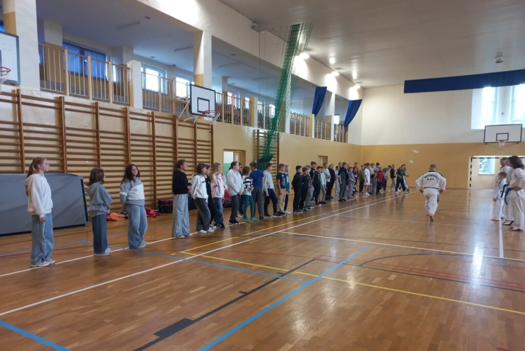 Europejski Dzień Sportu Szkolnego. Na zdjęciu trenerzy taekwondo pokazują uczniom ustawionym w szeregu elementy rozgrzewki: wykroki rozciągające.
