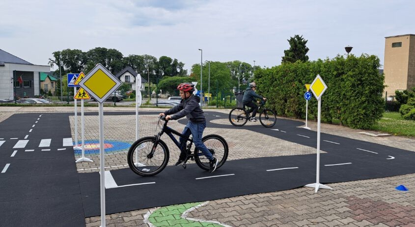 Odblaskowa Szkoła 2025. Na zdjęciu uczniowie poruszają się po Miasteczku Ruchu Drogowego. Na rowerach ćwiczą prawidłowe i bezpieczne poruszanie się po drodze.
