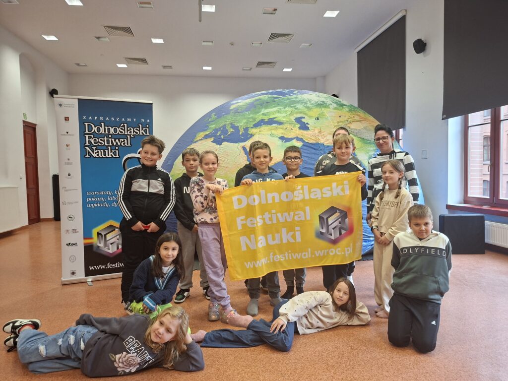 Zdjęcie przedstawia uczniów trzymających żółtą flagę z białym napisem Dolnośląski Festiwal Nauki. W tle sali jest Mobilne Planetatium, czyli kopuła kuli ziemskiej.