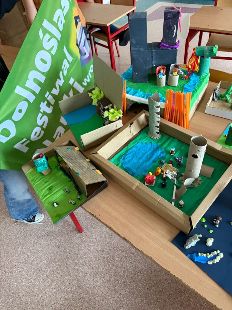 Przedstawia makiety Państwa Polskiego wykonane z kartonów, rolek oklejonych kolorowym papierem. W tle ławki oraz zielona flaga z białym napisem Dolnośląski Festiwal Nauki.