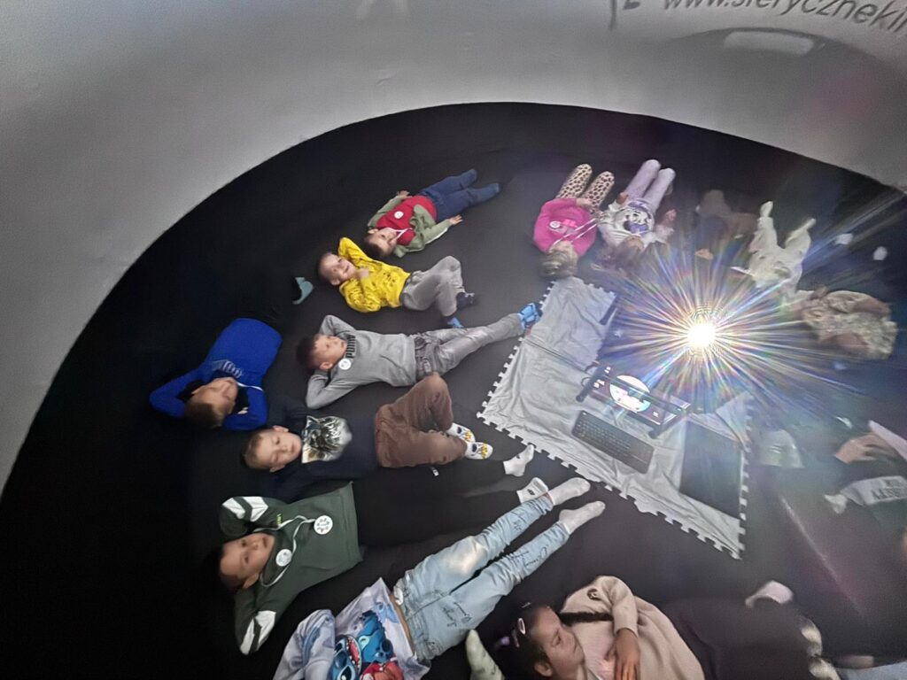 Zdjęcie przedstawia leżących w kole na czarnej podłodze uczniów, którzy obserwują w Mobilnym Planetarium film o kosmosie.