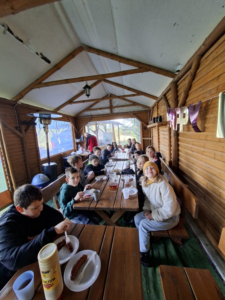 Zdjęcie przedstawia uczniów jedzących kiełbaski z ogniska w drewnianych pomieszczeniu. Na stołach znajdują się kubki z herbatą oraz chleb.