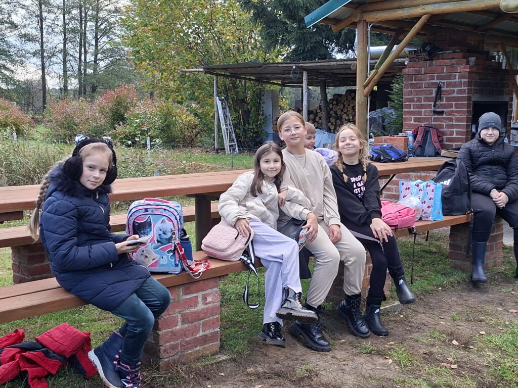 Zdjęcie przedstawia uczniów siedzących na ławce I odpoczywających po ognisku. W tle polana.