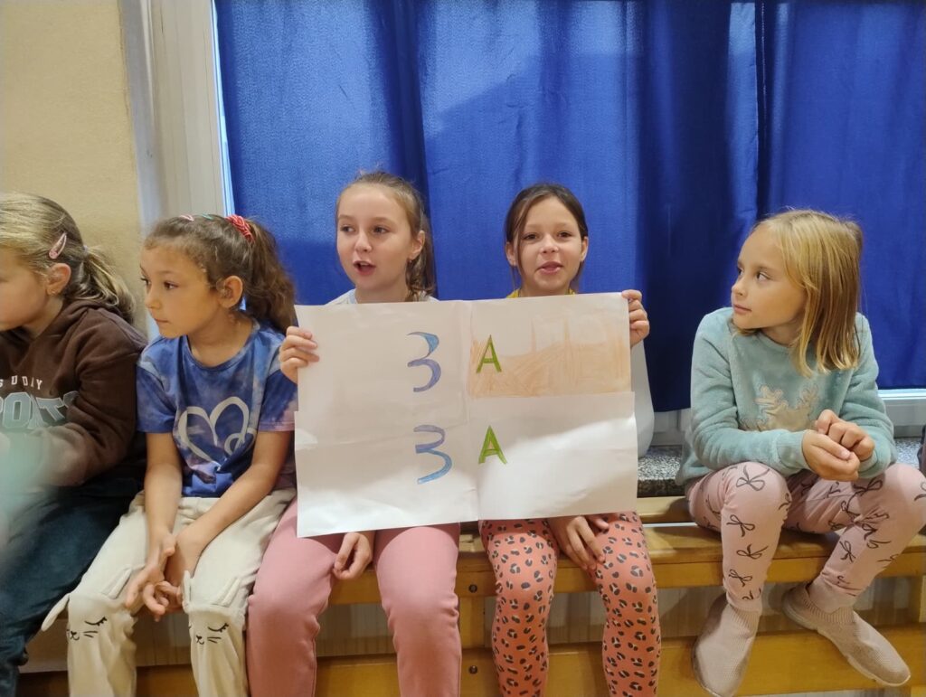 Dzień Chłopaka w klasach 1-3. Dziewczynki radośnie kibicują swojej klasie, trzymając transparent z napisem "3A!" i uśmiechając się do kamery.