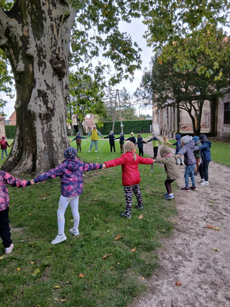 Wycieczka edukacyjna do Dalkowa. Na zdjęciu uczniowie trzymając się za ręce tworzą koło wokół wielkiego drzewa. Z prawej strony widać stary pałac.