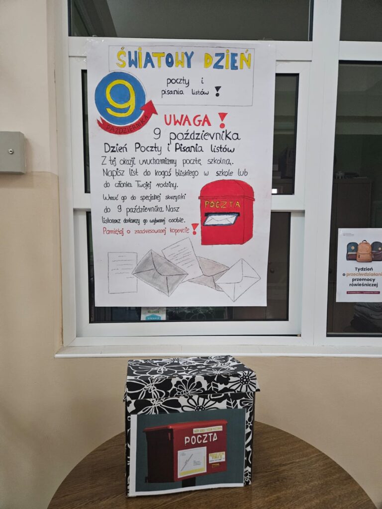 Ludzie Listy Piszą... Czyli Światowy Dzień Poczty W Naszej Szkole. Na zdjęciu plakat promujący akcję pisania listów 9 października. Przed plakatem stoi stolik, na którym umieszczono szkolną skrzynkę pocztową na listy uczniów.