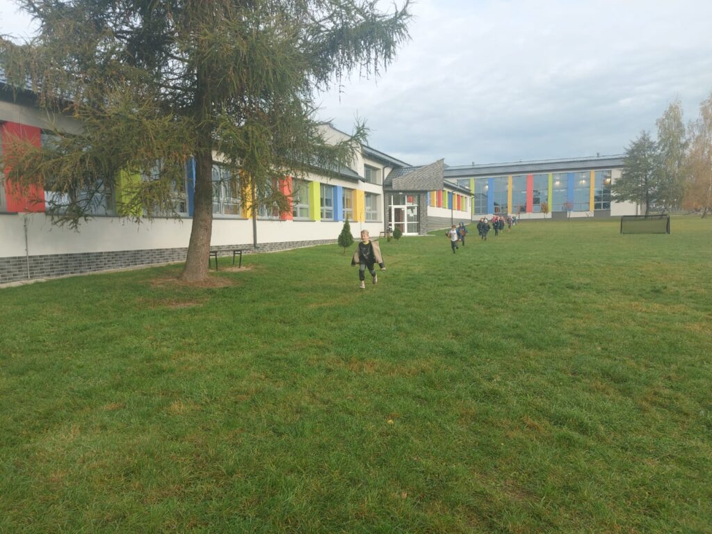 Uczniowie klasy pierwszej  biegną po wyznaczonej trasie na terenach zielonych wokół  szkoły. Po lewej stronie widać budynek szkoły.