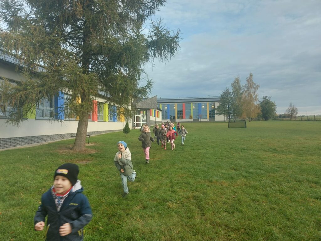 Uczniowie klasy pierwszej  b biegną po wyznaczonej trasie na terenach zielonych wokół  szkoły. Po lewej stronie widać budynek szkoły.