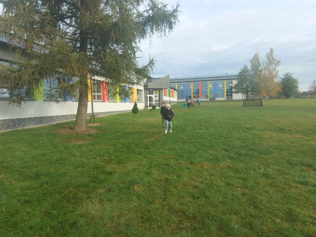 Uczniowie klasy pierwszej c  biegną po wyznaczonej trasie na terenach zielonych wokół  szkoły. Po lewej stronie widać budynek szkoły.