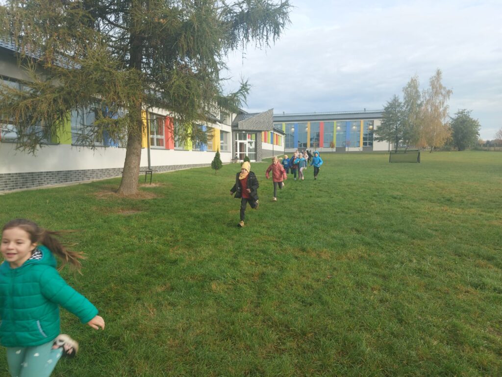 Uczniowie klasy trzeciej a  biegną po wyznaczonej trasie na terenach zielonych wokół  szkoły. Po lewej stronie widać budynek szkoły.