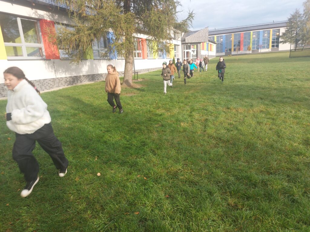 Uczniowie klasy drugiej a biegną po wyznaczonej trasie na terenach zielonych wokół  szkoły. Po lewej stronie widać budynek szkoły.