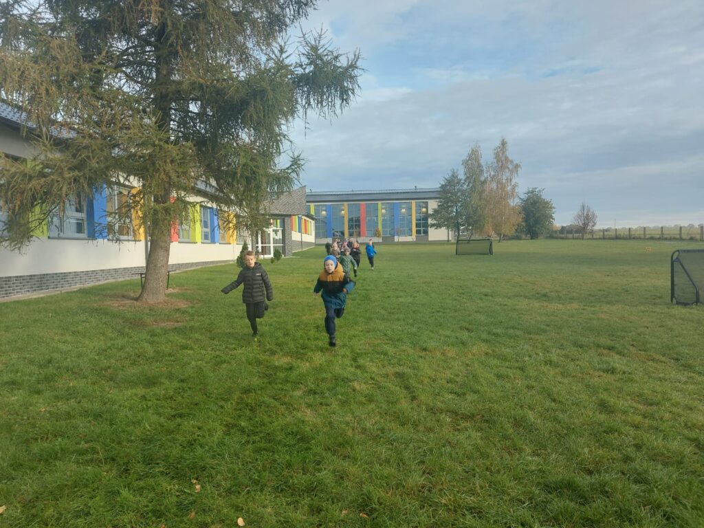 Uczniowie klasy drugiej b  biegną po wyznaczonej trasie na terenach zielonych wokół  szkoły. Po lewej stronie widać budynek szkoły.