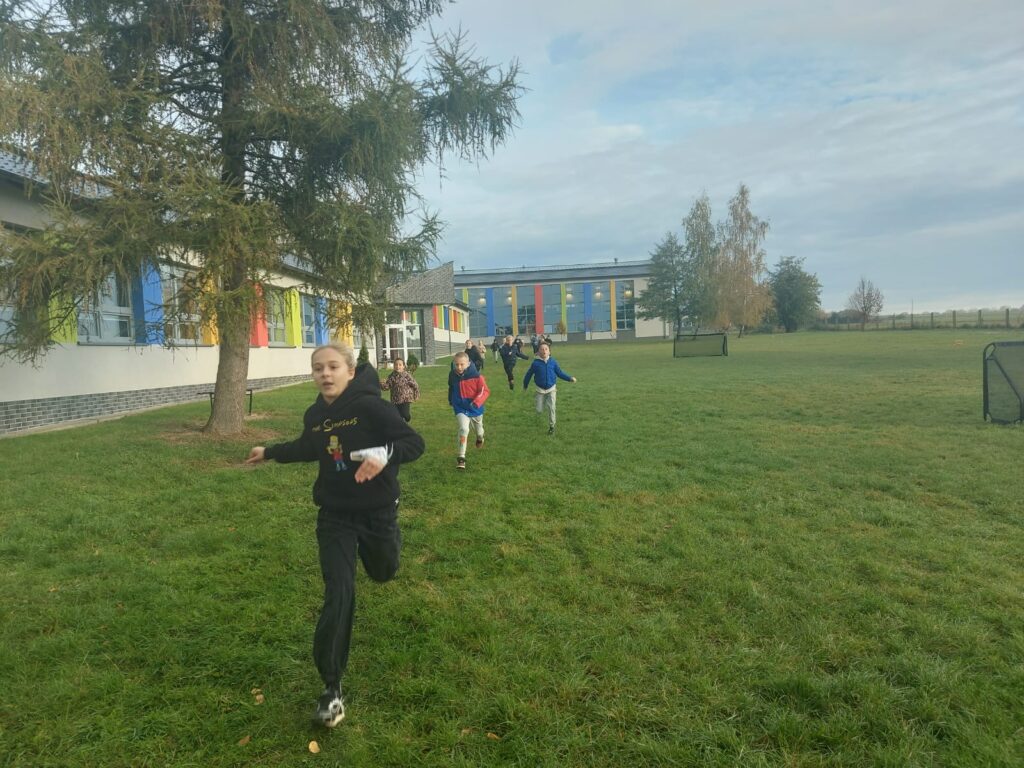 Uczniowie klasy trzeciej a  biegną po wyznaczonej trasie na terenach zielonych wokół  szkoły. Po lewej stronie widać budynek szkoły.