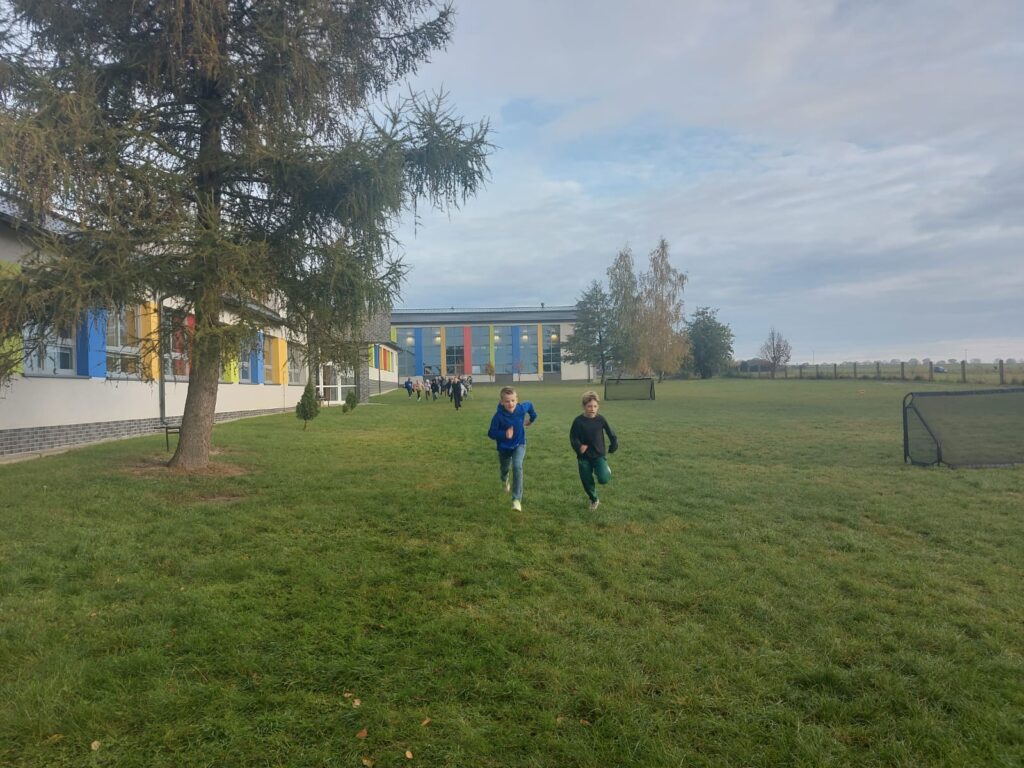 Uczniowie klasy trzeciej b  biegną po wyznaczonej trasie na terenach zielonych wokół  szkoły. Po lewej stronie widać budynek szkoły.