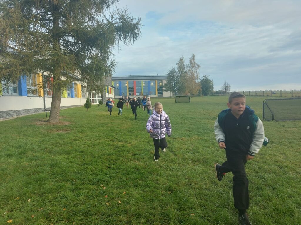 Uczniowie klasy trzeciej b  biegną po wyznaczonej trasie na terenach zielonych wokół  szkoły. Po lewej stronie widać budynek szkoły.