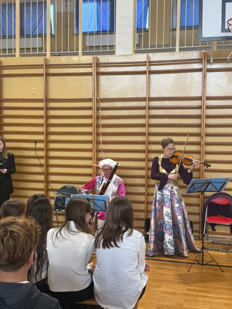 Koncert filharmonii. Na zdjęciu uczniowie klas 4-6 siedzą na ławeczkach na sali gimnastycznej ubrali na galowo, przed uczniami stoją muzycy: po środku pan w białej peruce i różowym stroju gra na  klarnecie, po prawej stronie pani ubrana w długą suknię gra na skrzypcach.