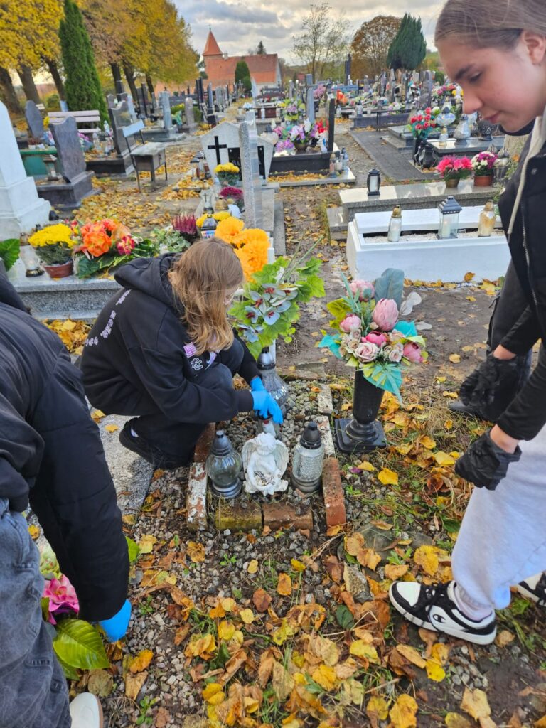 Ocalić od zapomnienia, czyli sprzątamy opuszczone groby. Na zdjęciu mały grób. Z lewej strony dwie wolontariuszki. Jedna stoi, druga porządkuje grób. Z prawej strony wolontariuszka