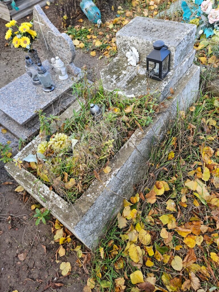 Ocalić od zapomnienia, czyli sprzątamy opuszczone groby. Na zdjęciu duży, stary, murowany nagrobek. Cały porośnięty trawą i chwastami. Na środku stare kwiaty i znicze.