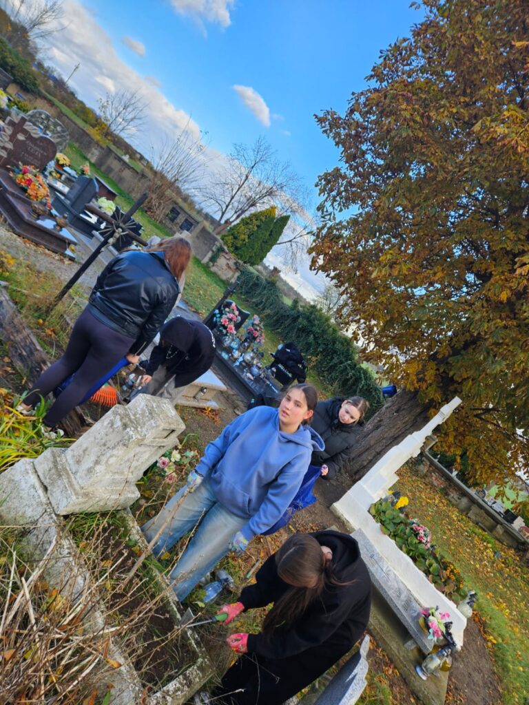 Ocalić od zapomnienia, czyli sprzątamy opuszczone groby. Na zdjęciu dwie wolontariuszki porządkujące duży grób. Z lewej stoi wolontariuszka w niebieskiej bluzie, z prawej wolontariuszka usuwająca trawę. W tle po lewej stronie widać inną grupę sprzątających wolontariuszy.